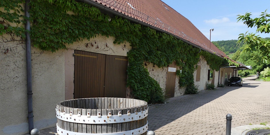 Ein mit Efeu bewachsenes Gebäude mit einem roten Dach steht neben einer gepflasterten Straße. Vor dem Gebäude befindet sich ein großer Holztrog. Im Hintergrund sind Bäume und blauer Himmel zu sehen.