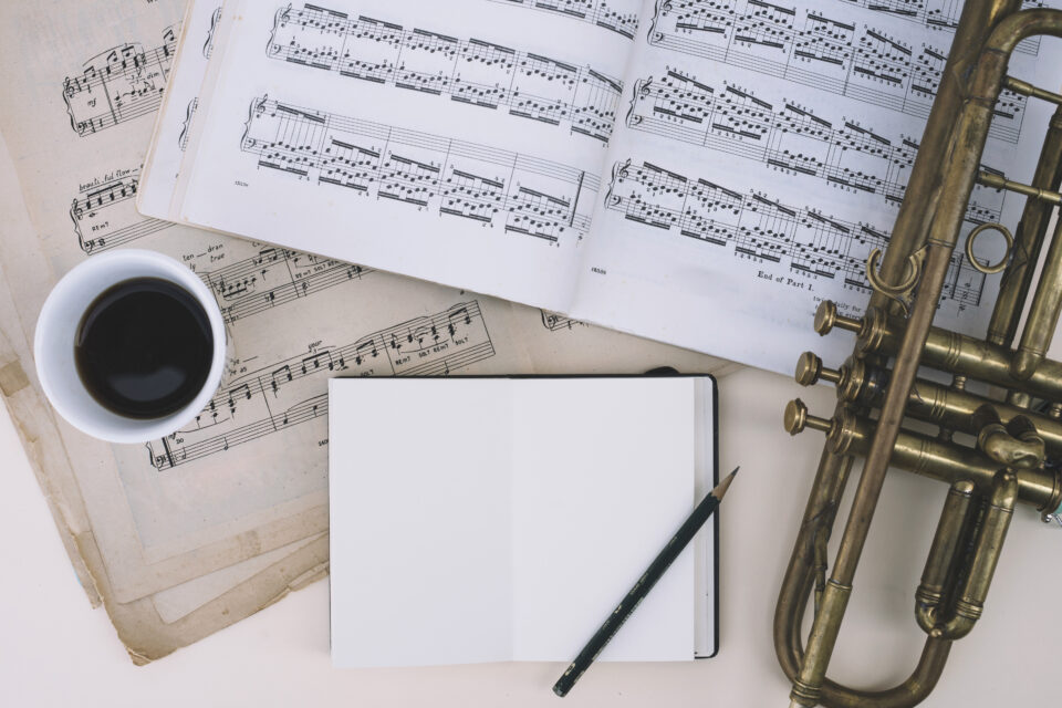 Auf dem Bild sind ein Notizbuch, ein Bleistift, eine Tasse Kaffee, Musiknoten und eine Trompete angeordnet. Die Notenblätter zeigen musikalische Notationen, während das Notizbuch unbeschrieben auf einem Tisch liegt.