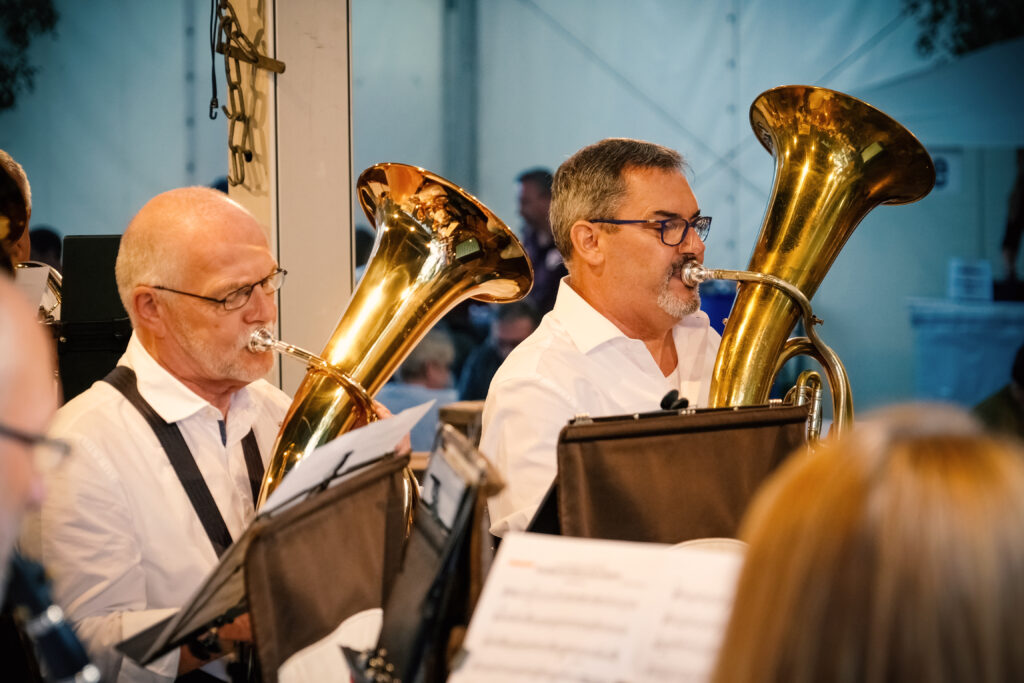 Zwei Musiker spielen Tuba in einer Blaskapelle. Die Männer tragen weiße Hemden und sind umgeben von Notenständern. Im Hintergrund sind weitere Musiker und eine Veranstaltung zu erkennen. Die Szenerie vermittelt eine festliche Atmosphäre.