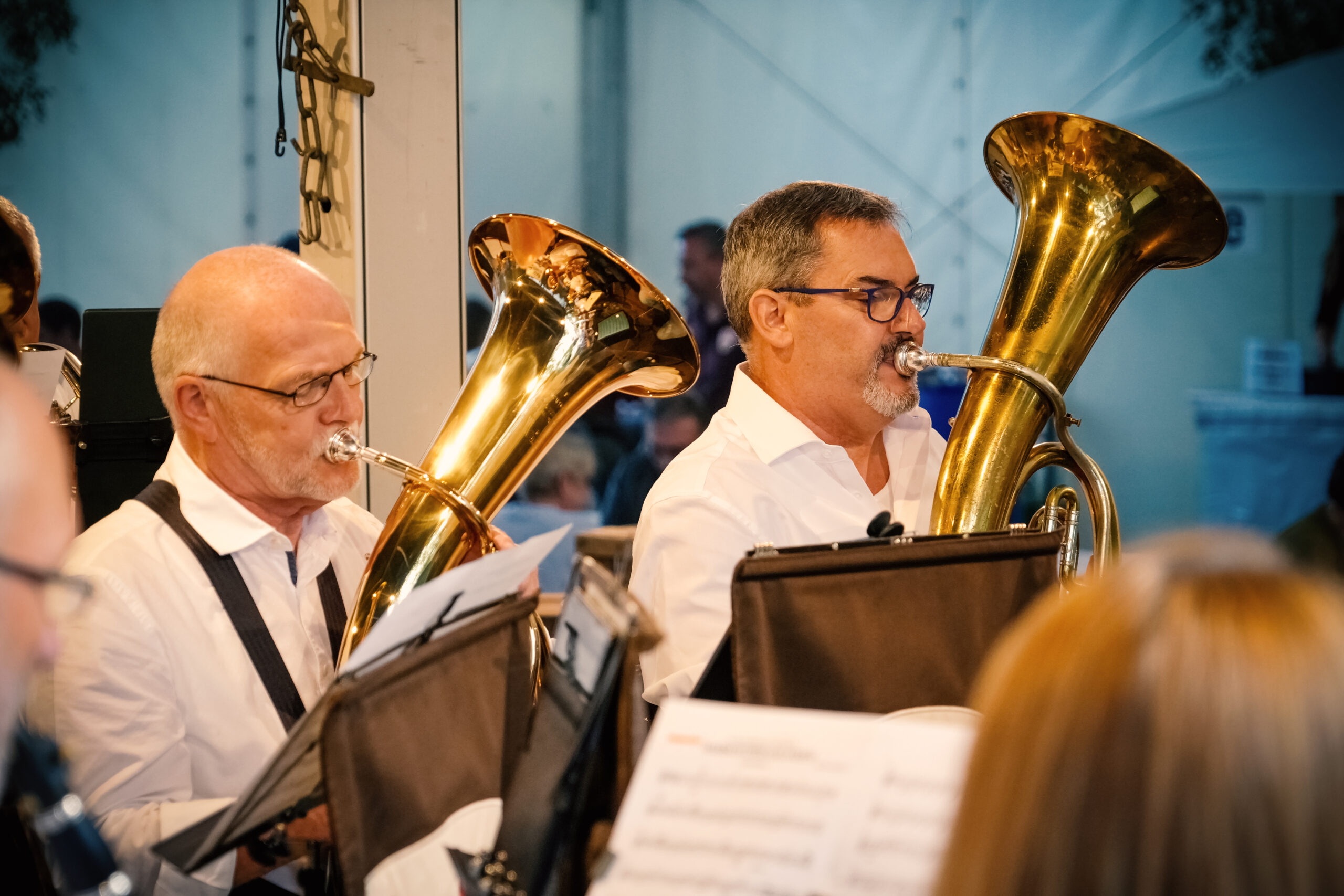 Zwei Musiker spielen Tuba in einer Blaskapelle. Die Männer tragen weiße Hemden und sind umgeben von Notenständern. Im Hintergrund sind weitere Musiker und eine Veranstaltung zu erkennen. Die Szenerie vermittelt eine festliche Atmosphäre.