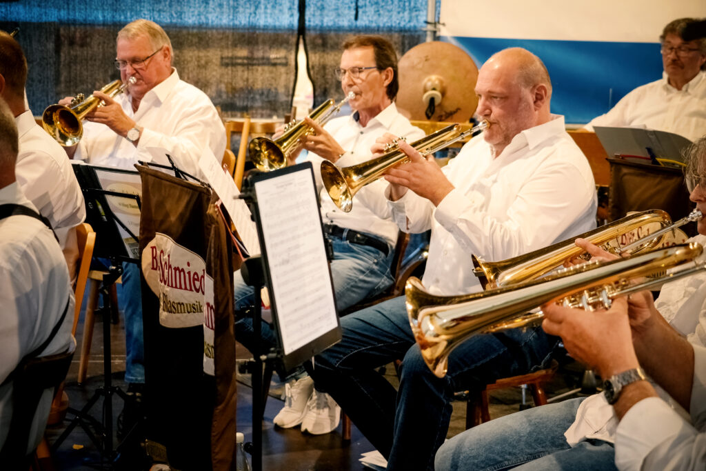 A group of musicians play trumpets in a wind orchestra. The musicians wear white shirts and sit at desks with sheet music. Various musical instruments are visible in the background. The atmosphere is lively and communal.   