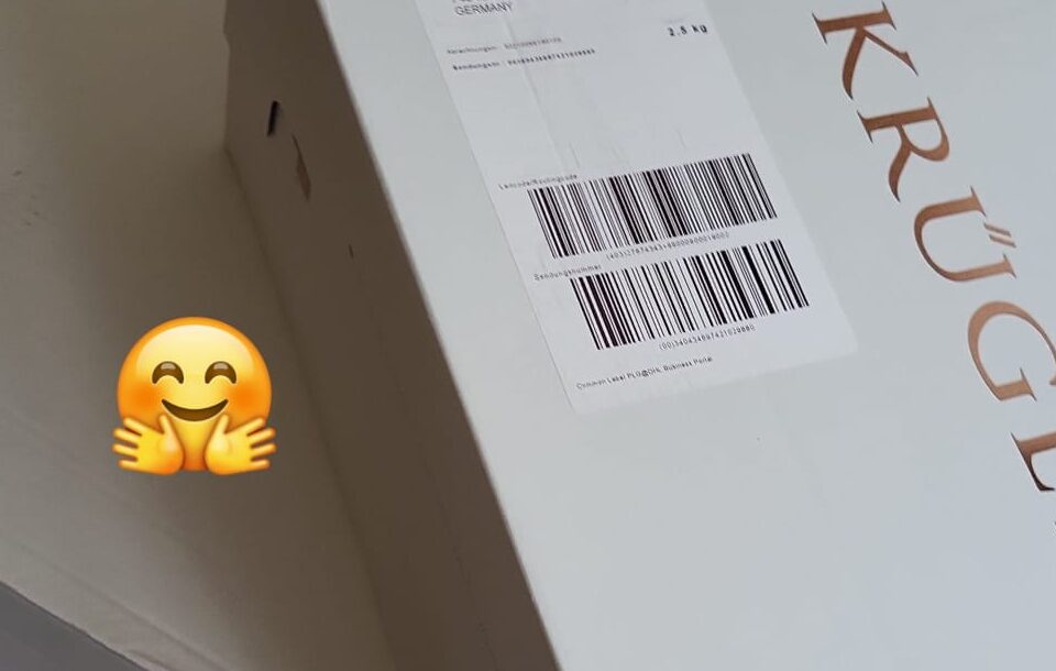 Eine weiße Versandbox mit dem Aufdruck "Krüger" und einem Barcode ist auf einem Tisch platziert. Neben der Box befindet sich ein Emoji, das ein lächelndes Gesicht mit offenen Armen zeigt.
