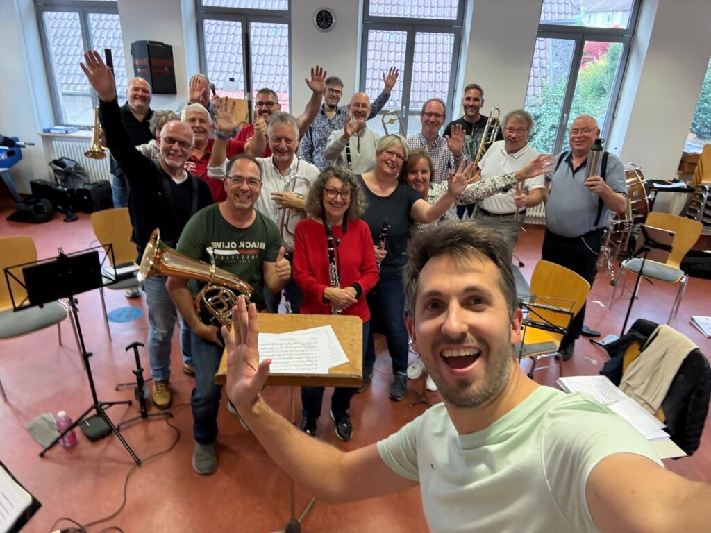A group of about twenty people stand in a room and smile at the camera. They all raise their hands. In the foreground is a man taking a selfie. Music stands and instruments can be seen in the background.   