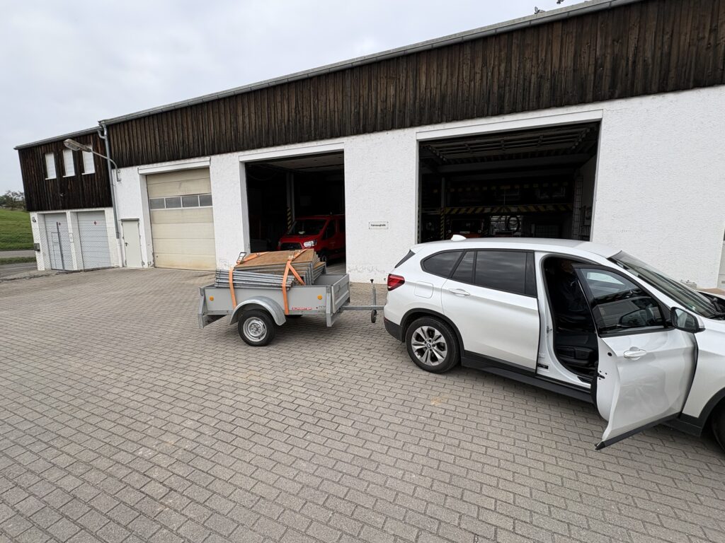 Ein weißer SUV steht auf einem gepflasterten Platz vor einer großen Lagerhalle. Der Kofferraum ist geöffnet, und ein Anhänger mit einem Holzstapel ist hinter dem Auto angekuppelt. Die Hallentore sind teilweise geöffnet, der Himmel ist bewölkt.