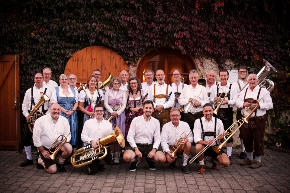 A group of people in traditional dress stand in front of a wall covered with plants. They are holding wind instruments such as tubas, trumpets, and trombones.